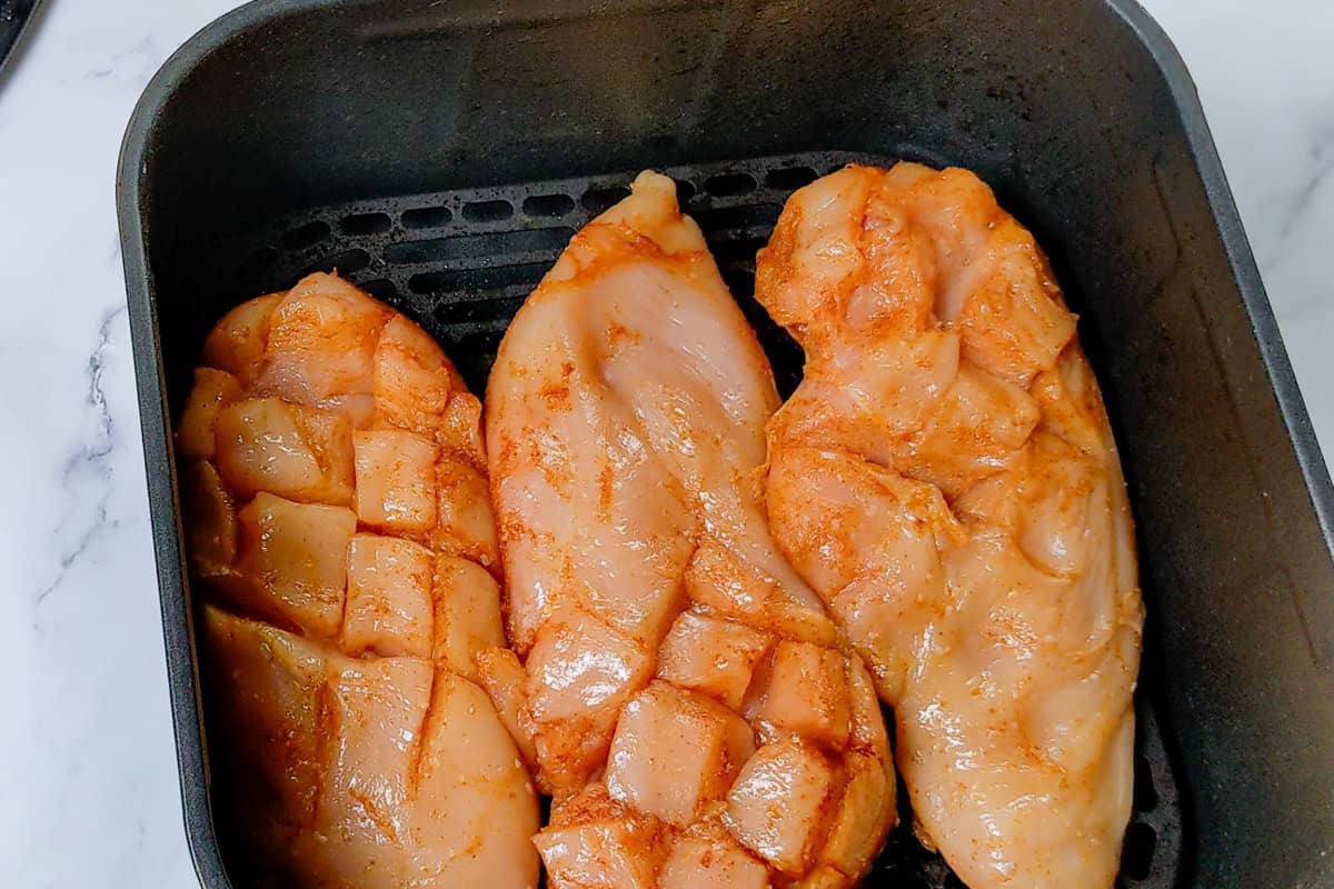 Raw seasoned chicken breasts with shallow diamond cuts before air frying in an air fryer basket.
