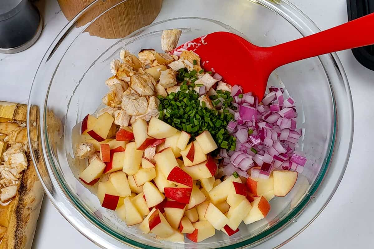 diced apple, chopped chives, chopped red onions and diced cooked chicken in a pyrex glass mixing bowl with a red silicone spatula.