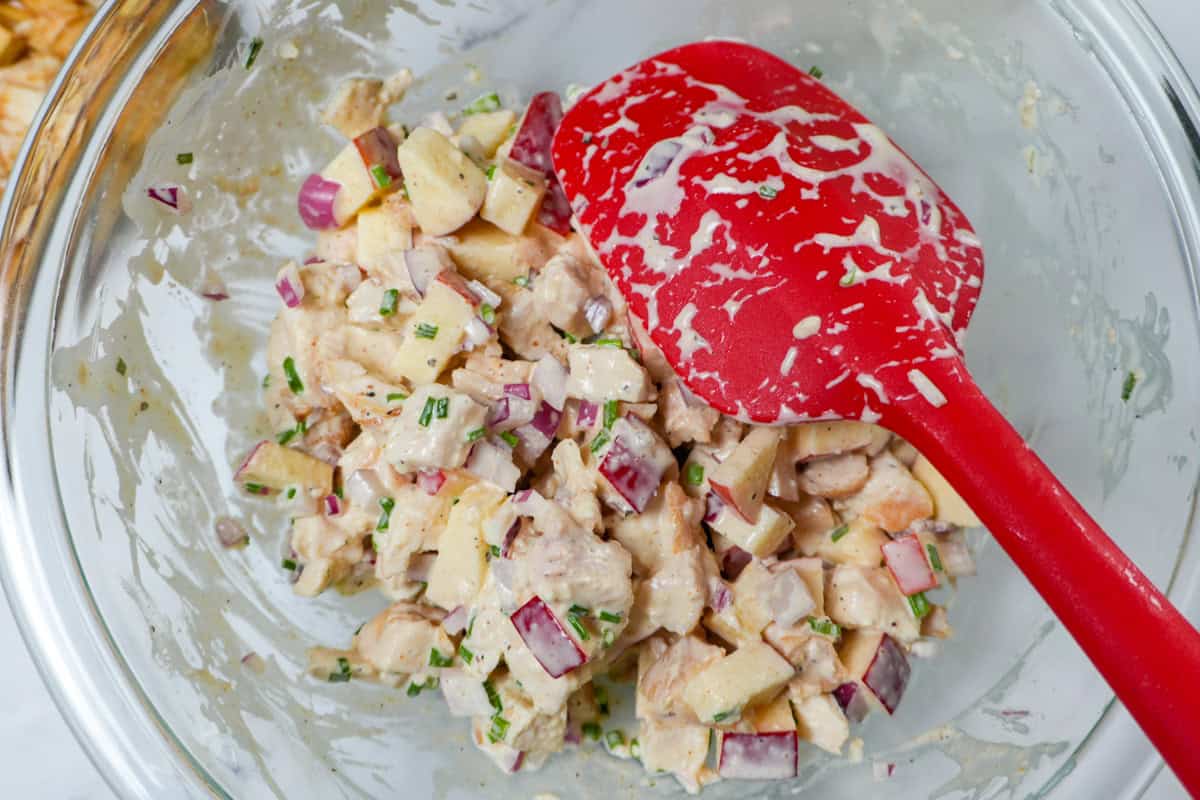 Small diced chicken and apples folded into creamy dressing with red onion and chives in a mixing bowl.