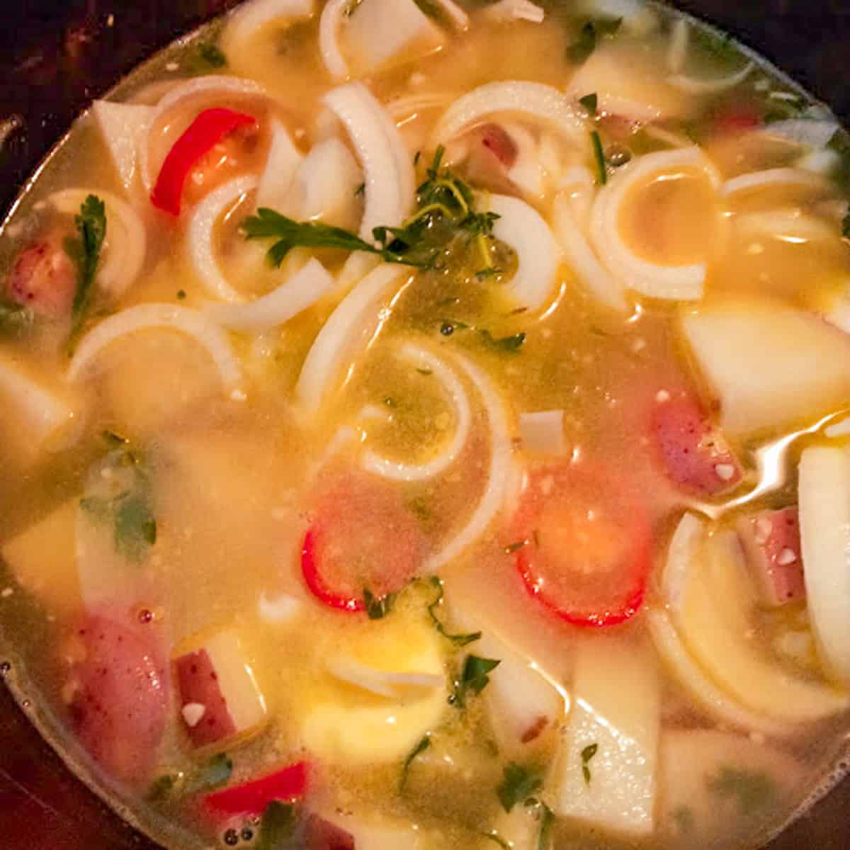 Potatoes, onions, herbs, and red peppers simmering in a pot of broth.