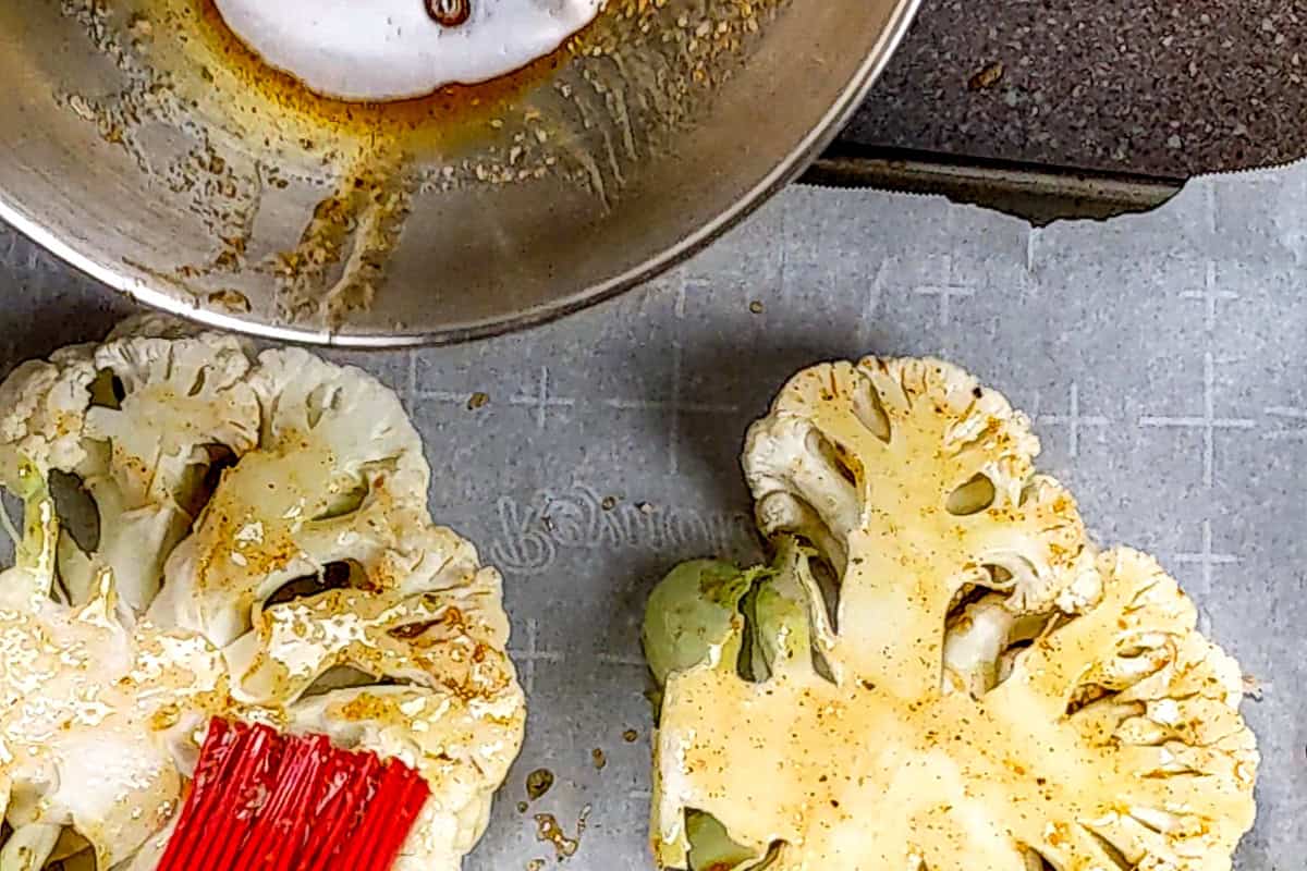 Cauliflower steaks brushed with wet rub seasoning on parchment paper on a sheet pan.