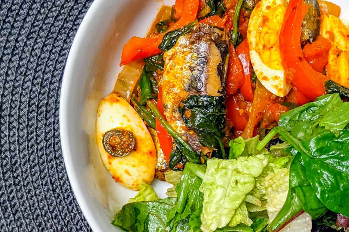 Smoked sardines with harissa, spinach, red bell peppers, tomatoes, and boiled eggs in a white wide salad bowl with salad on the side on a black weaved table mat.