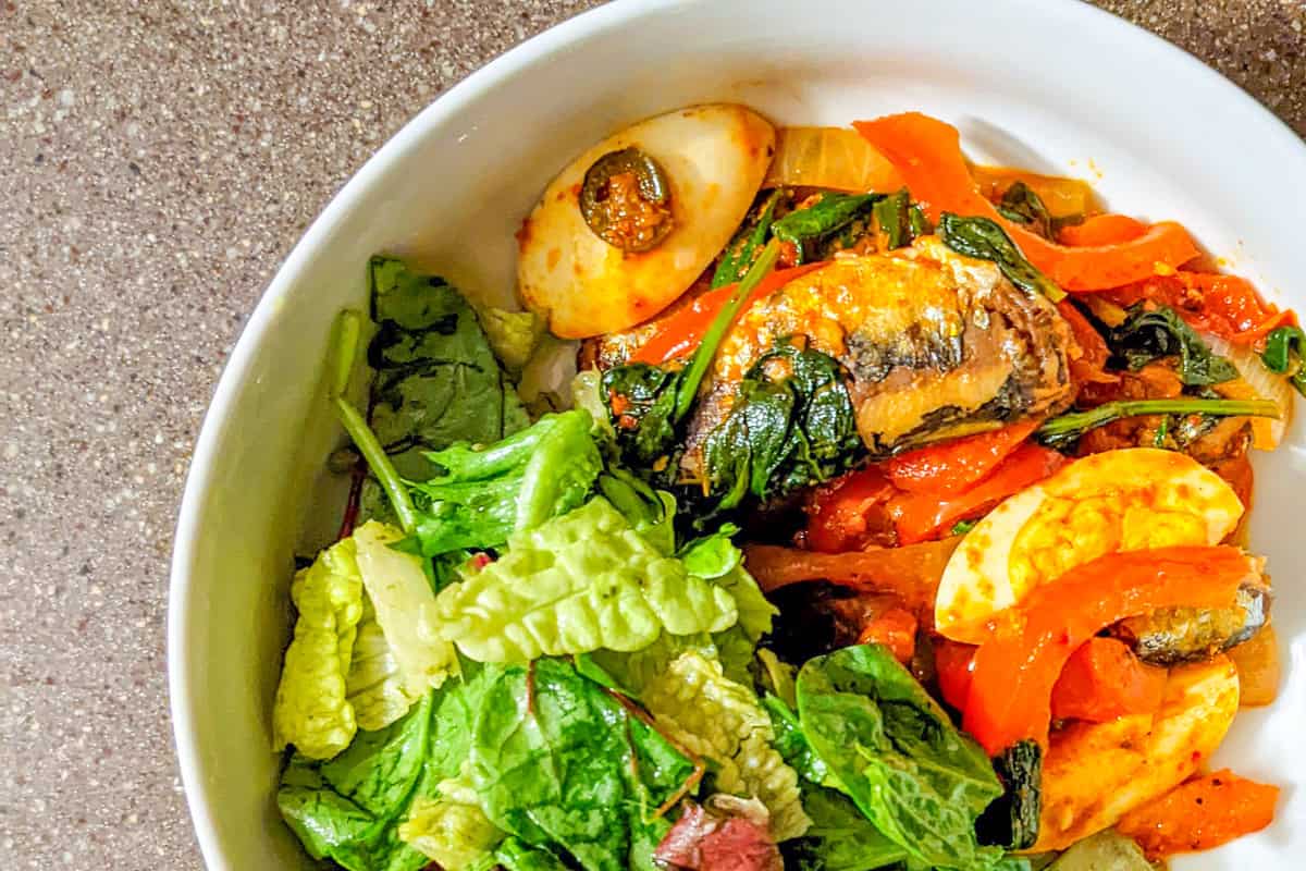 Smoked sardines with harissa, spinach, red bell peppers, tomatoes, and boiled eggs in a white wide salad bowl with salad on the side.