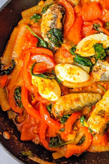 Smoked sardines with harissa, spinach, red bell peppers, tomatoes, and boiled eggs in a skillet.