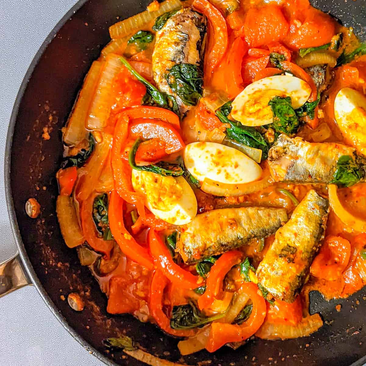 Smoked sardines with harissa, spinach, red bell peppers, tomatoes, and boiled eggs in a skillet.