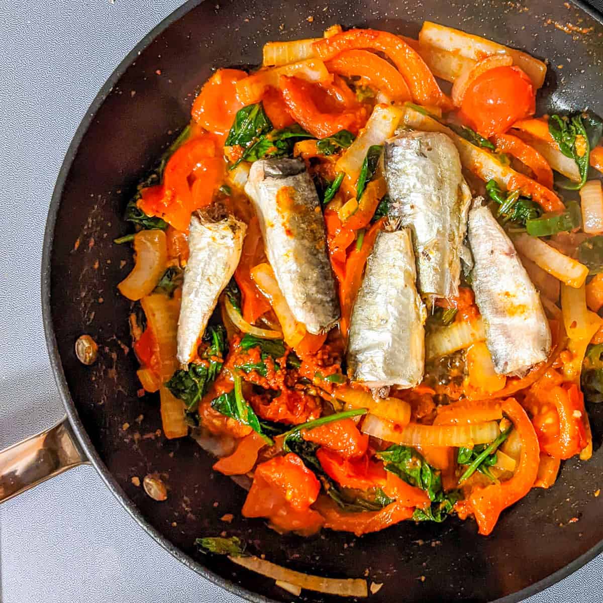 Smoked sardines on top of a harissa sauce with spinach, red bell peppers, tomatoes, and in a skillet.