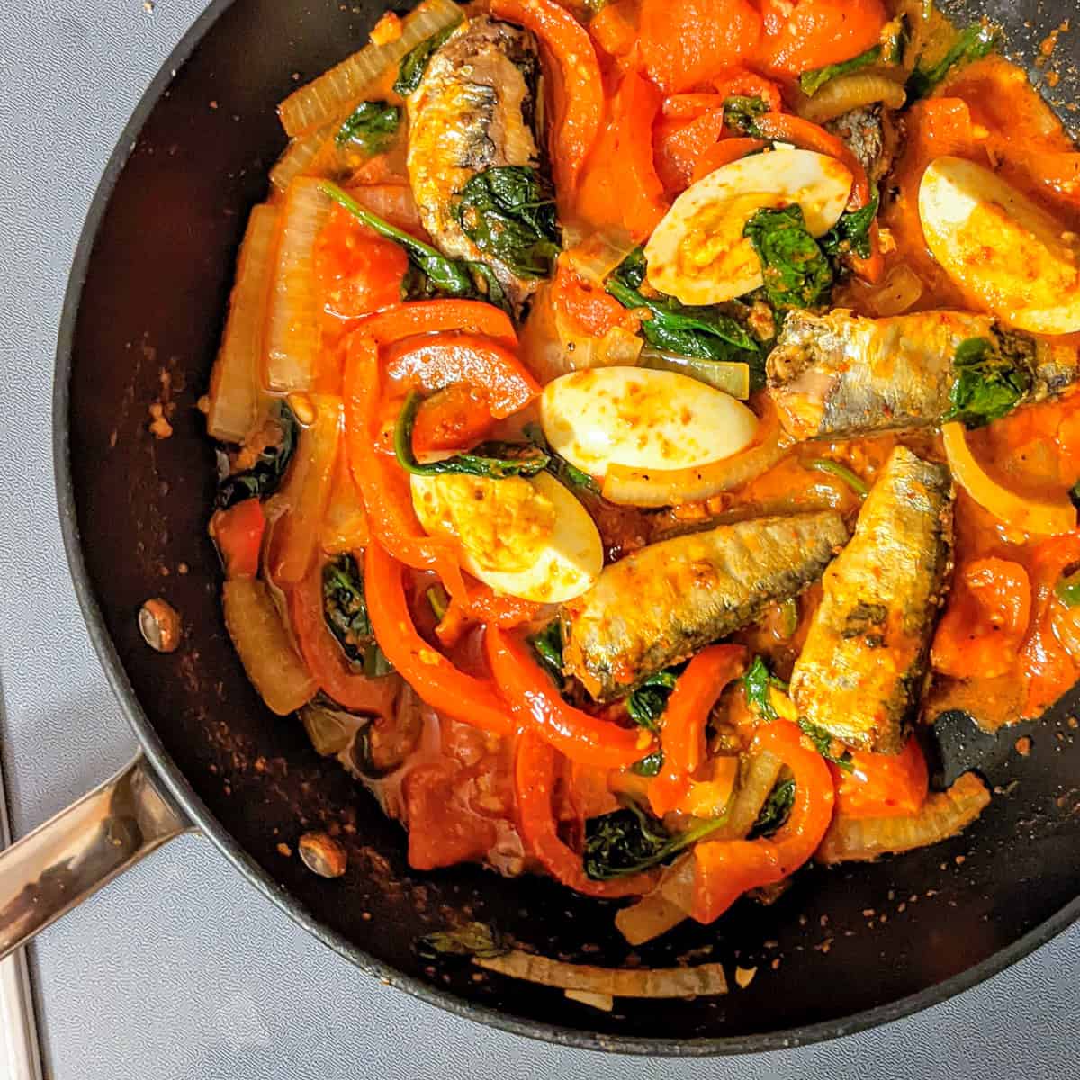 Smoked sardines with harissa, spinach, red bell peppers, tomatoes, and boiled eggs in a skillet.