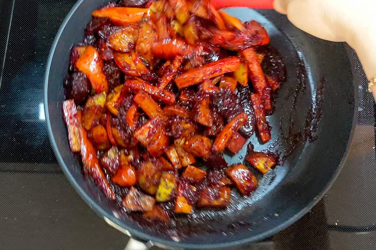 Roasted vegetables being gently folded into a red beet sauce in a non-stick fry pan.